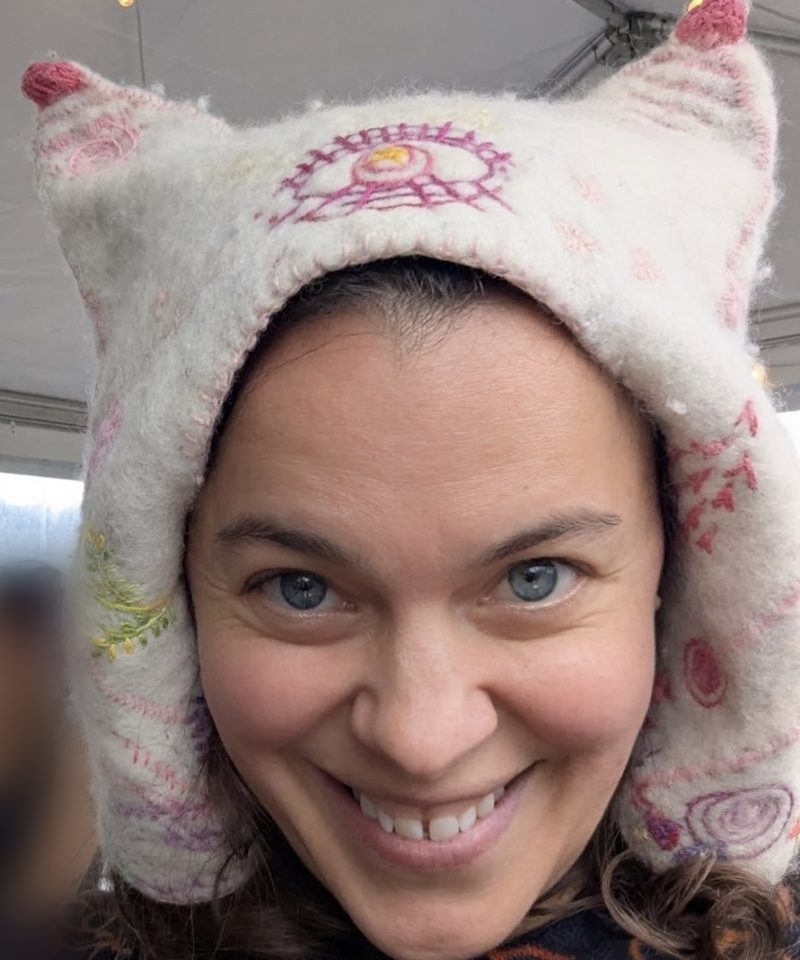 Elizabeth smiling in a white wool cat-ear sauna hat with pink embroidery